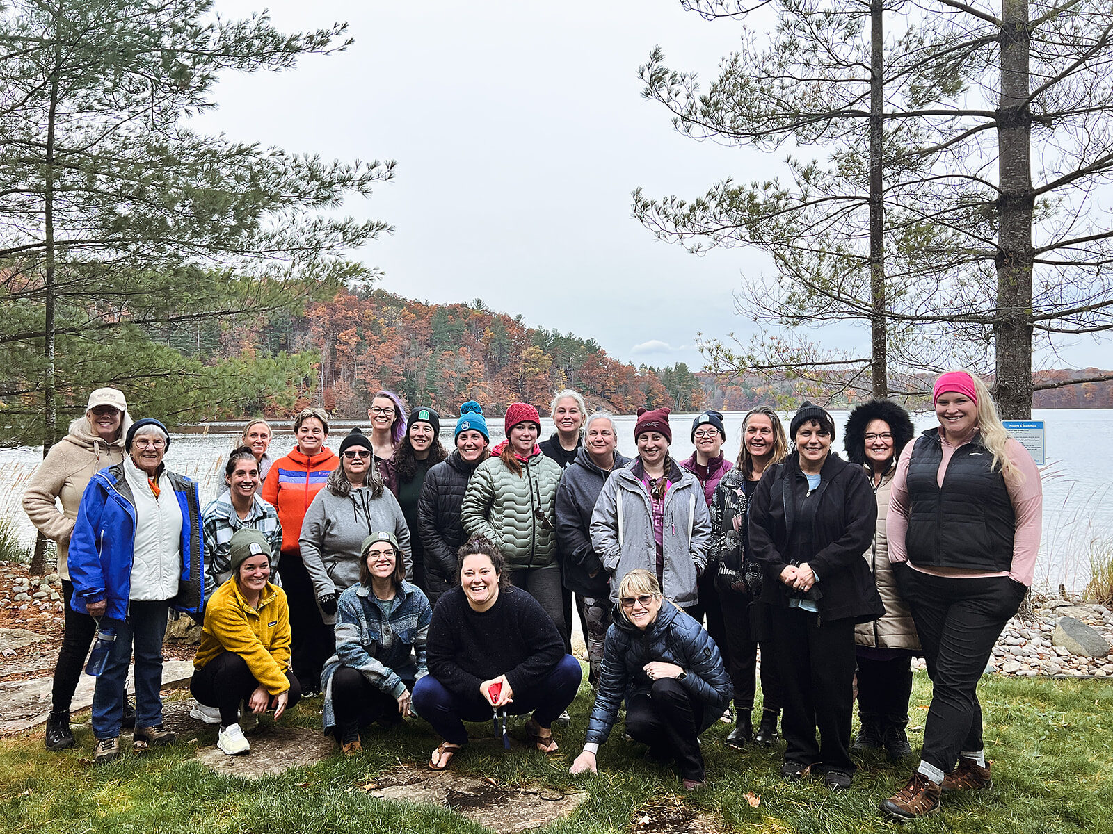 A large group of women standing together on the shore of a lake in autumn, posed for a group photograph.