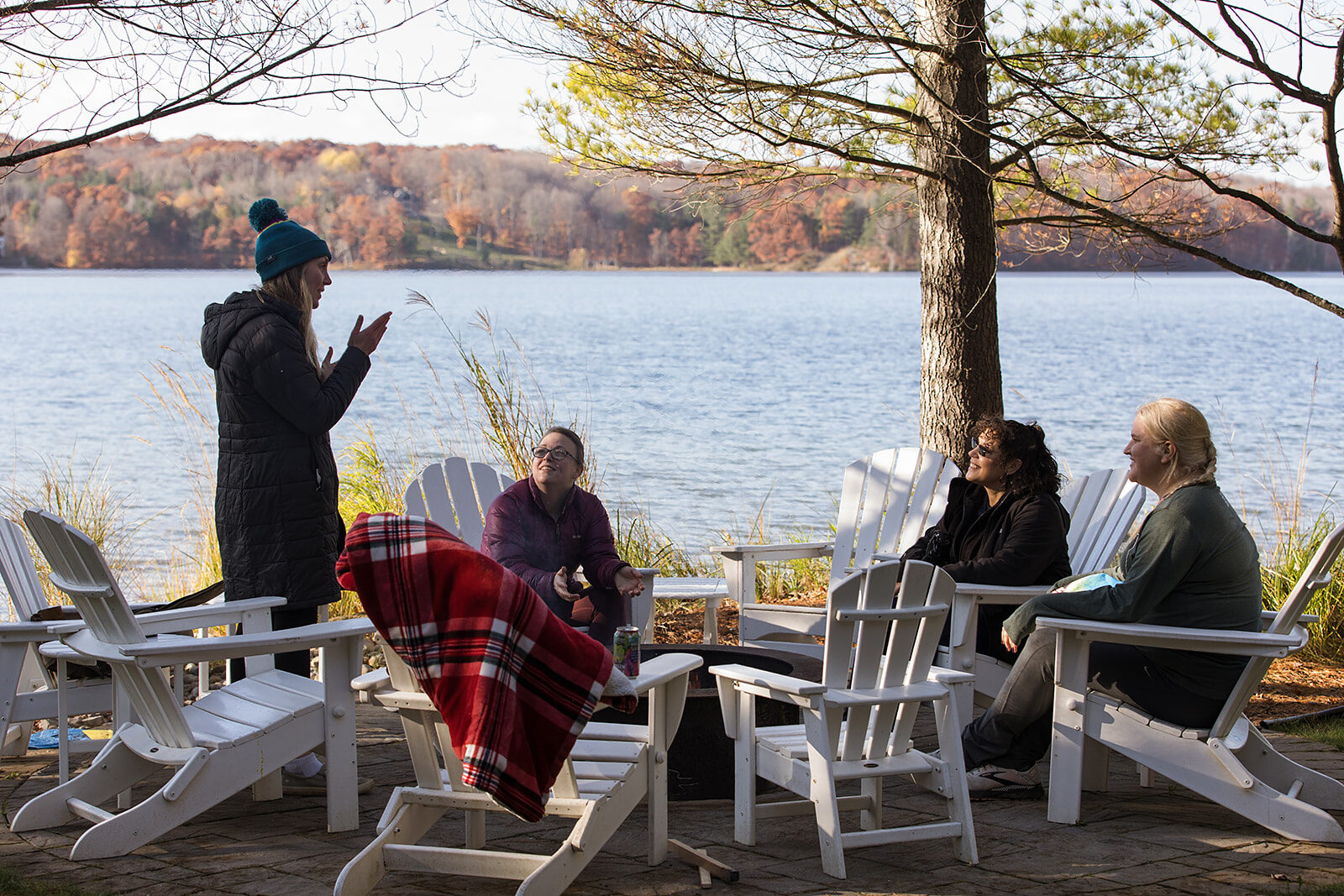 People sitting in a circle of chairs around an outdoor fire pit beside a lake, one person standing and talking.