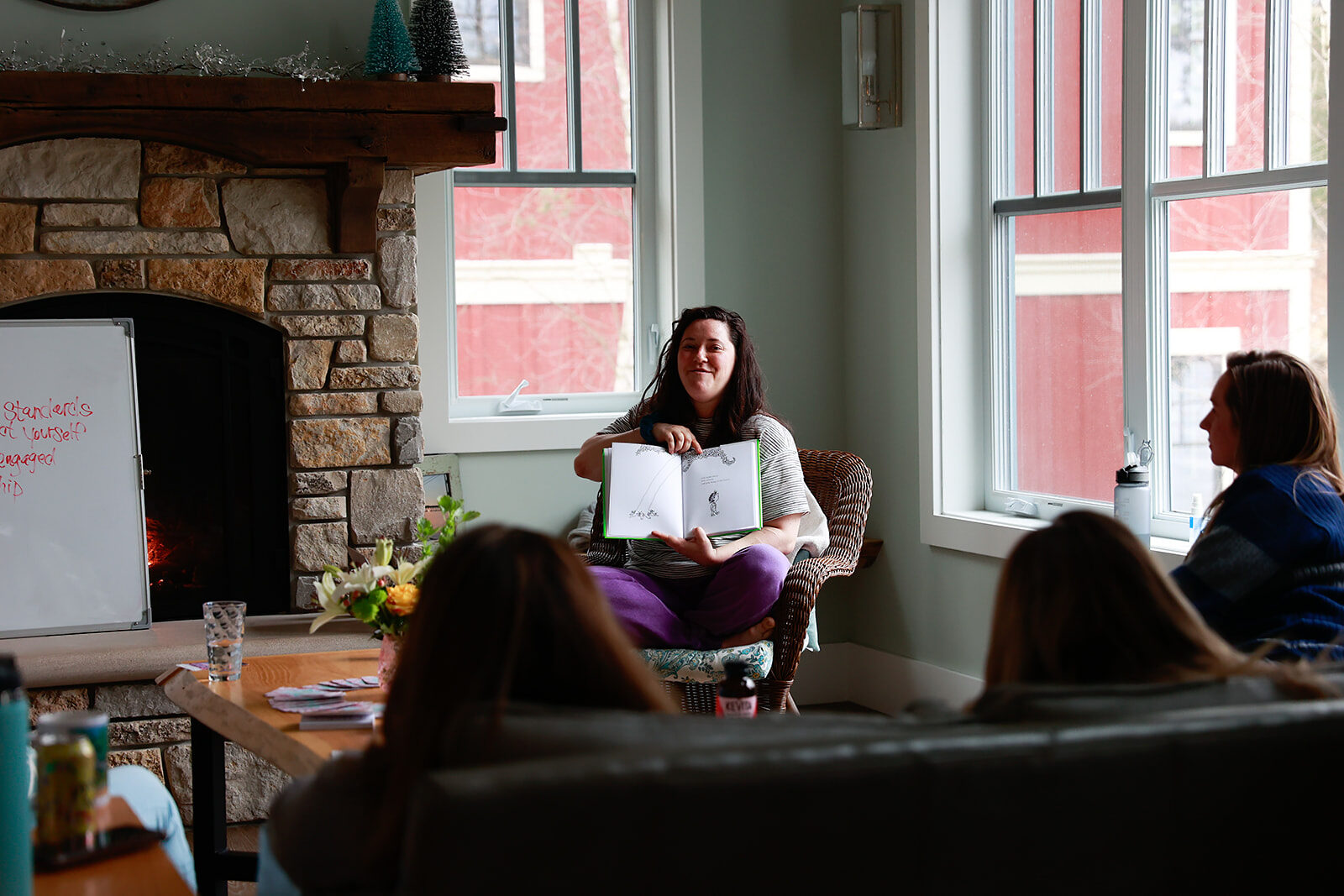 A woman presenting from an armchair with notes in her hand while a group listens, a fire lit behind them.
